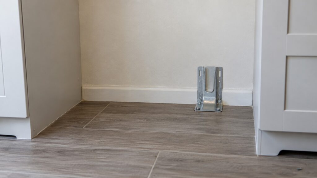 A metal mounting bracket fixed to a floor beside the rear wall of an empty range space, seen from a low angle, no text or numbers visible