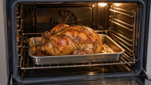 A roasted whole chicken with golden skin on a low-sided roasting pan inside a lit oven, no text or numbers visible