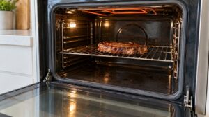 A built-in wall oven with its door open and a steak resting on a rack below the upper heating element